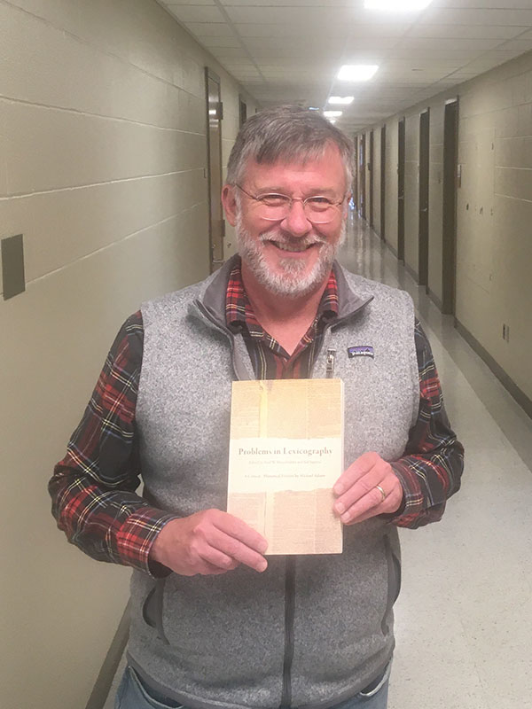 A photo of Professor Michael Adams, who holds a copy of his book and poses in a hallway in Ballantine Hall.
