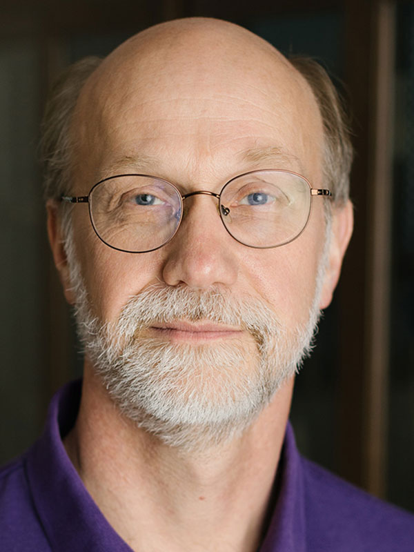 A headshot of Professor Ken de Jong, who wears a blue collared shirt and poses against a dark background.