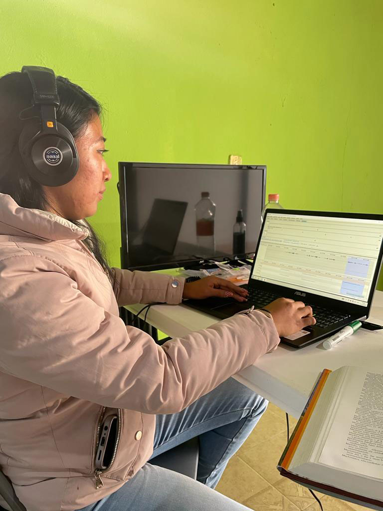 A woman works at a laptop computer. She wears a white coat and headphones.