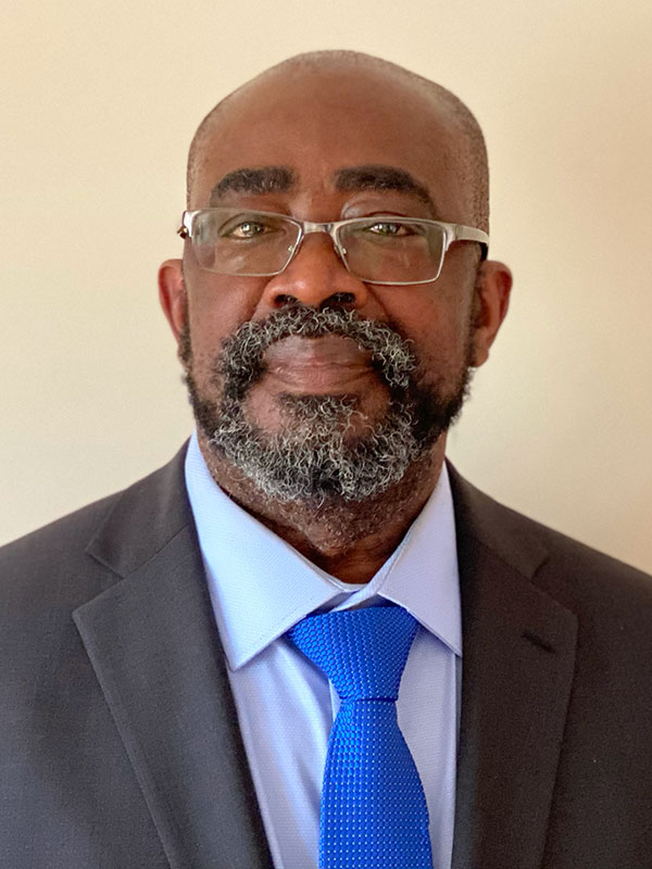 A headshot of Professor Samuel Obeng, who wears a dark suit, blue shirt, and necktie, and poses against a beige background.