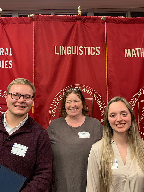 Three people pose in front of the Linguistics department banner at graduation.
